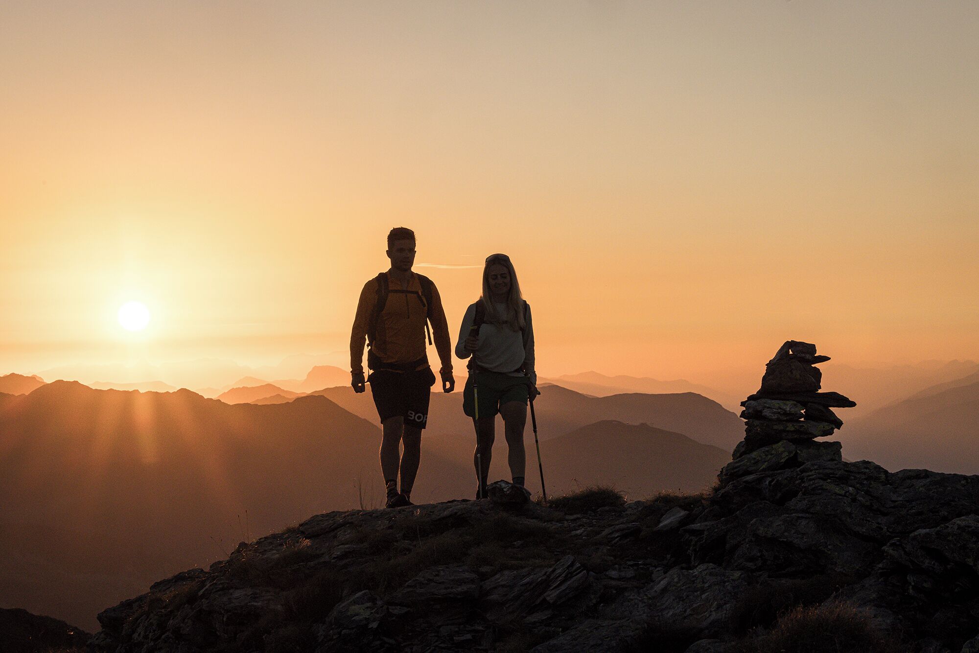 Zwei Wanderer auf einem Berg, im Hintergrund ein Sonnenuntergang.