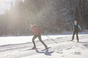 Duas pessoas esquiando de fundo em uma trilha nevada, com árvores ao fundo.