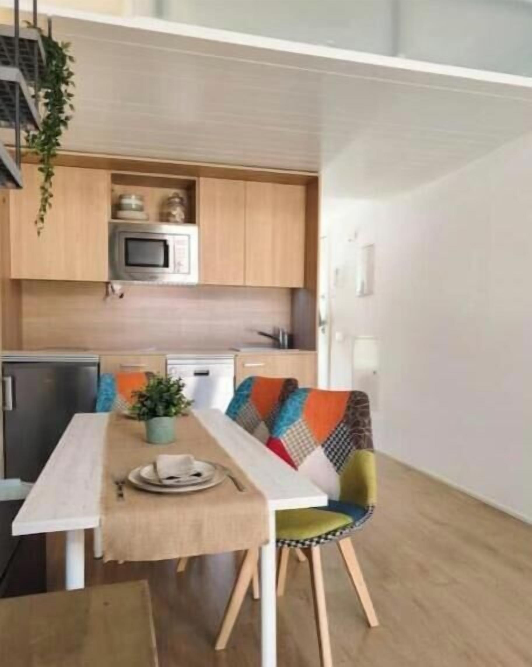 Een compacte keuken met een magnetron, een kleine koelkast en een vaatwasser. Er staat een eettafel met een bankje en een potplant op de tafel.