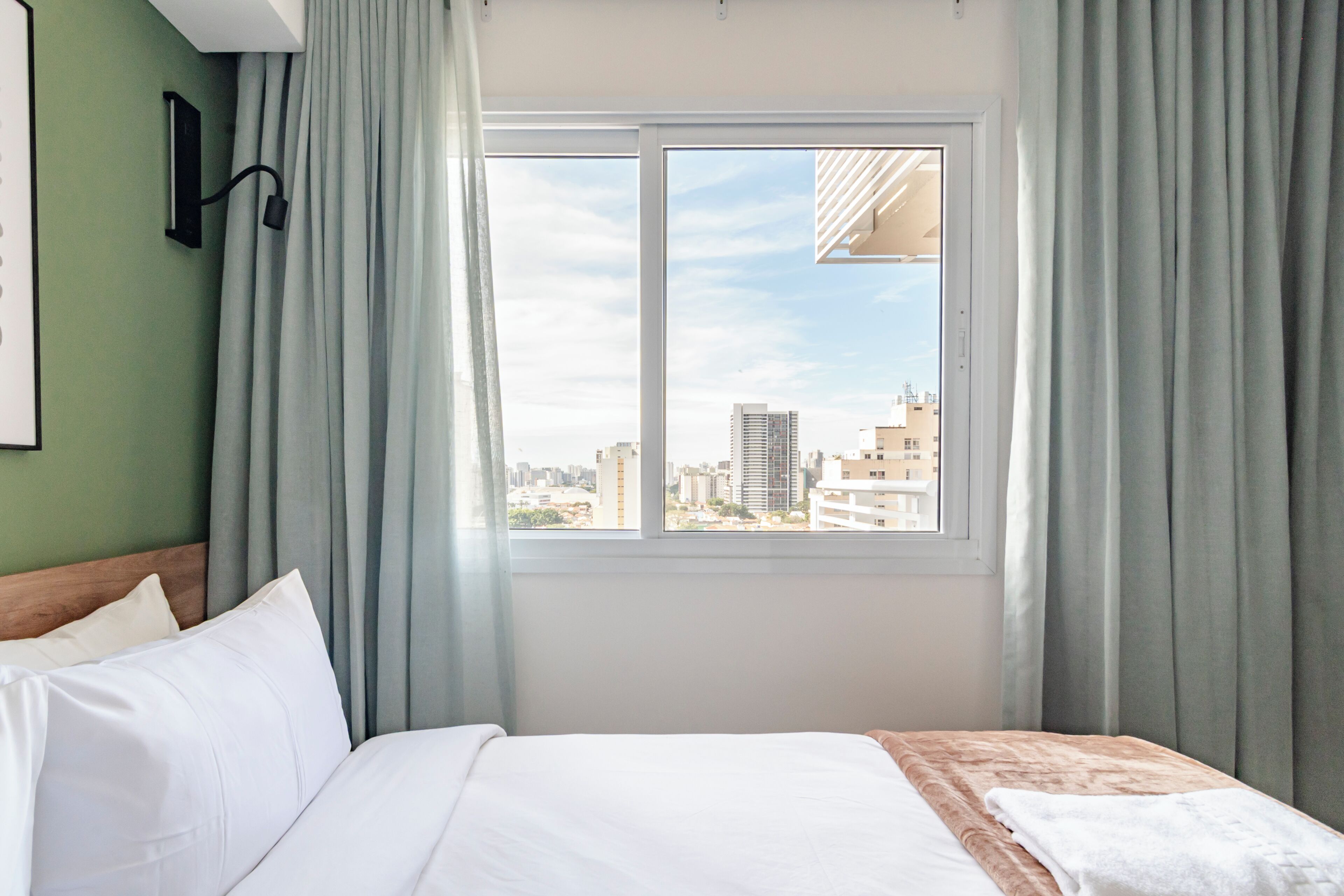 A hotel room with a bed, a window with curtains, a view of the cityscape, and a wall-mounted light fixture.