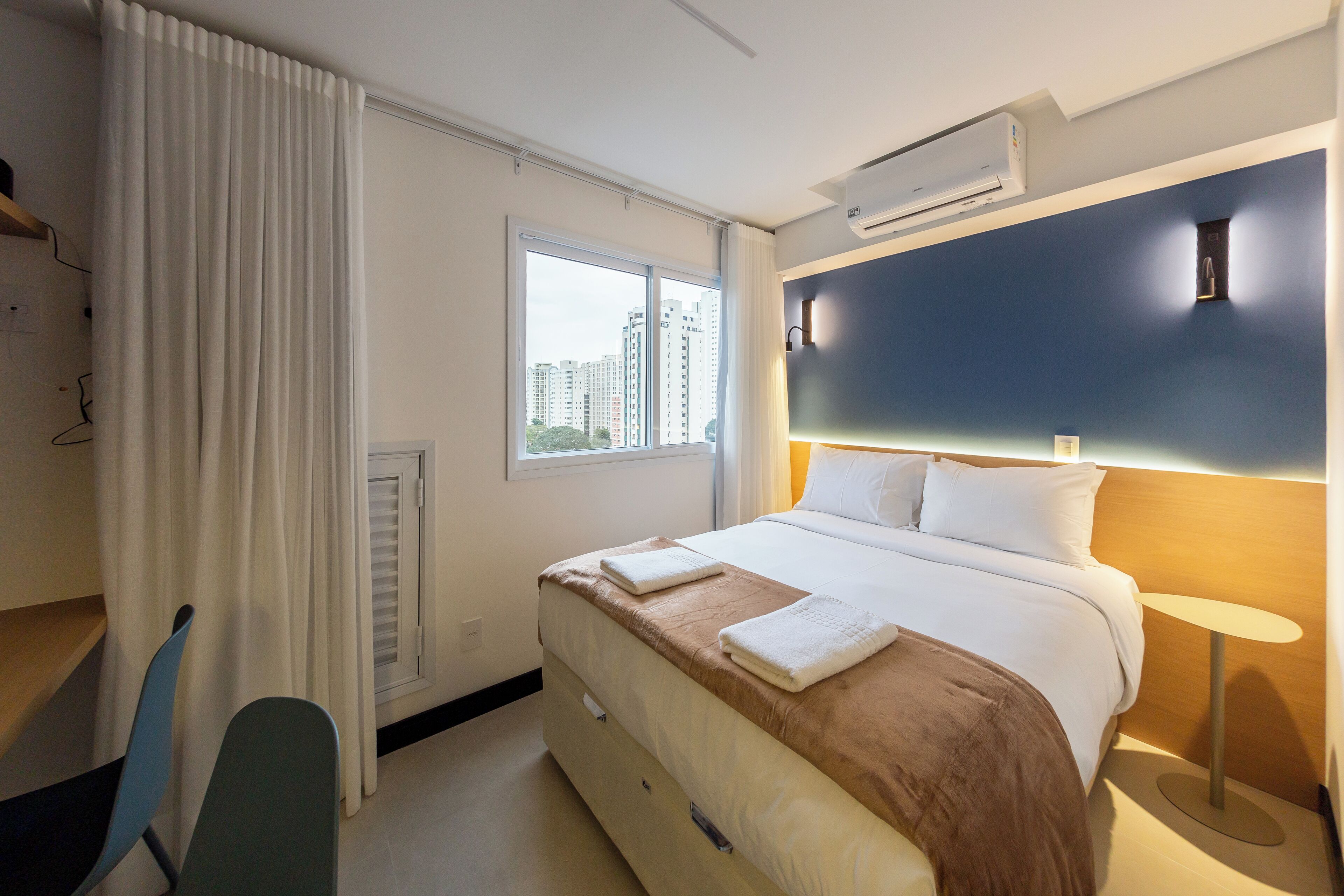 A hotel room with a bed, a desk, a chair, an air conditioning unit, a window, and a view of buildings.