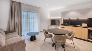 Una sala de estar moderna con sofá, una mesa redonda y sillas, una zona de cocina con televisor y un amplio ventanal con cortinas.