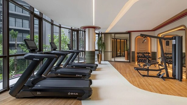Un gimnasio moderno con cintas de correr, máquinas de pesas y un amplio ventanal con vista a un edificio.