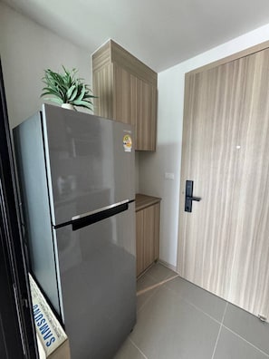 A compact kitchen with a stainless steel refrigerator, a wooden cabinet, and a door with a handle. - The Base • 1BR • Skyline View (Choeng Thale)