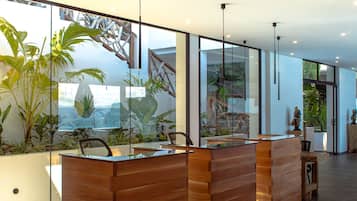 A reception desk with a wooden finish, glass panels, and a view of indoor plants and a mountainous landscape.