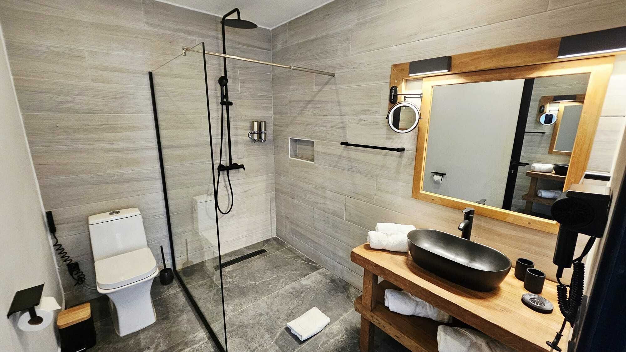 A modern bathroom with a glass-enclosed shower, a white toilet, a wooden vanity with a black sink, and a large mirror.