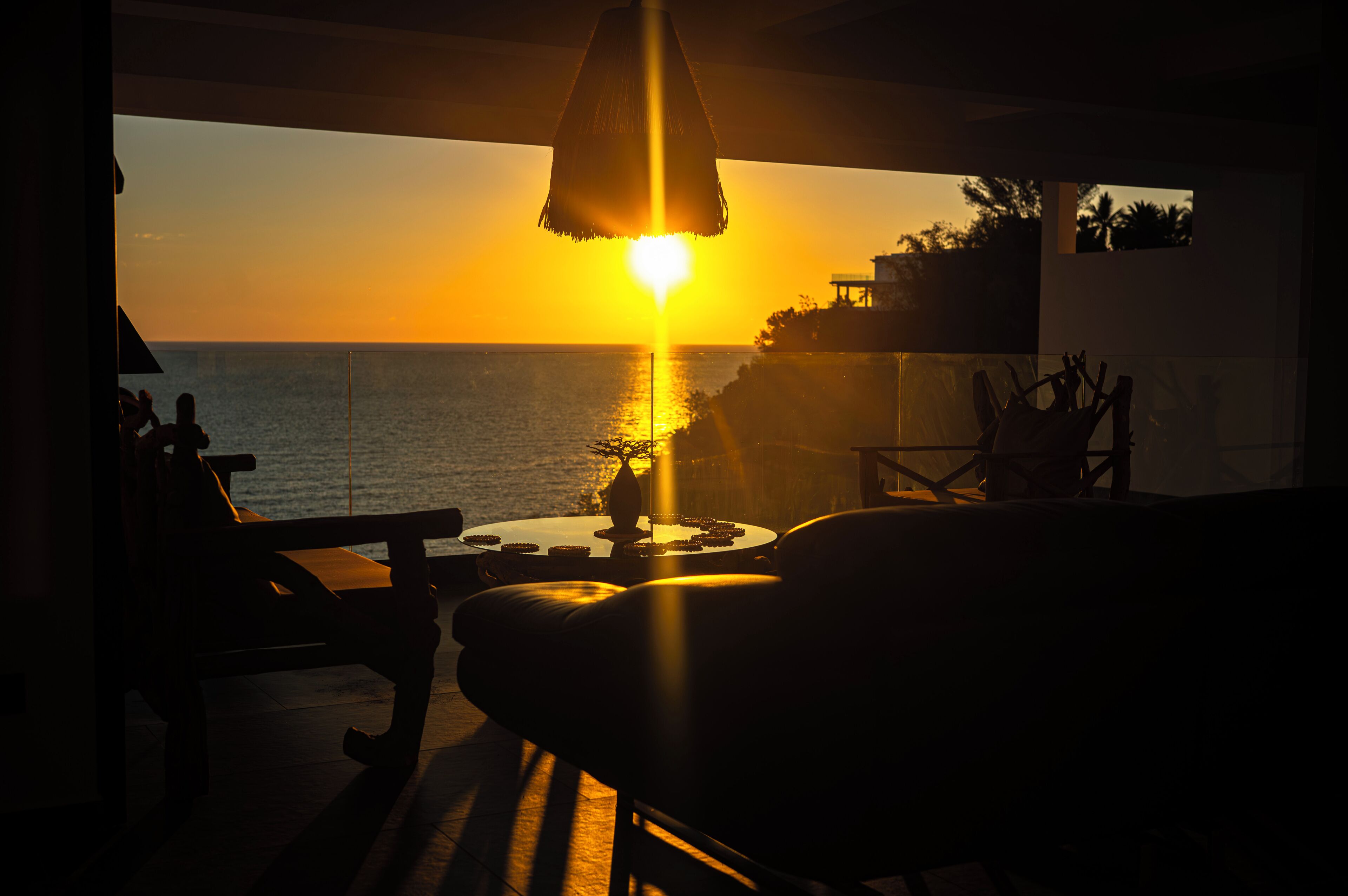 A sunset view from a balcony with a table and chairs, overlooking the sea.