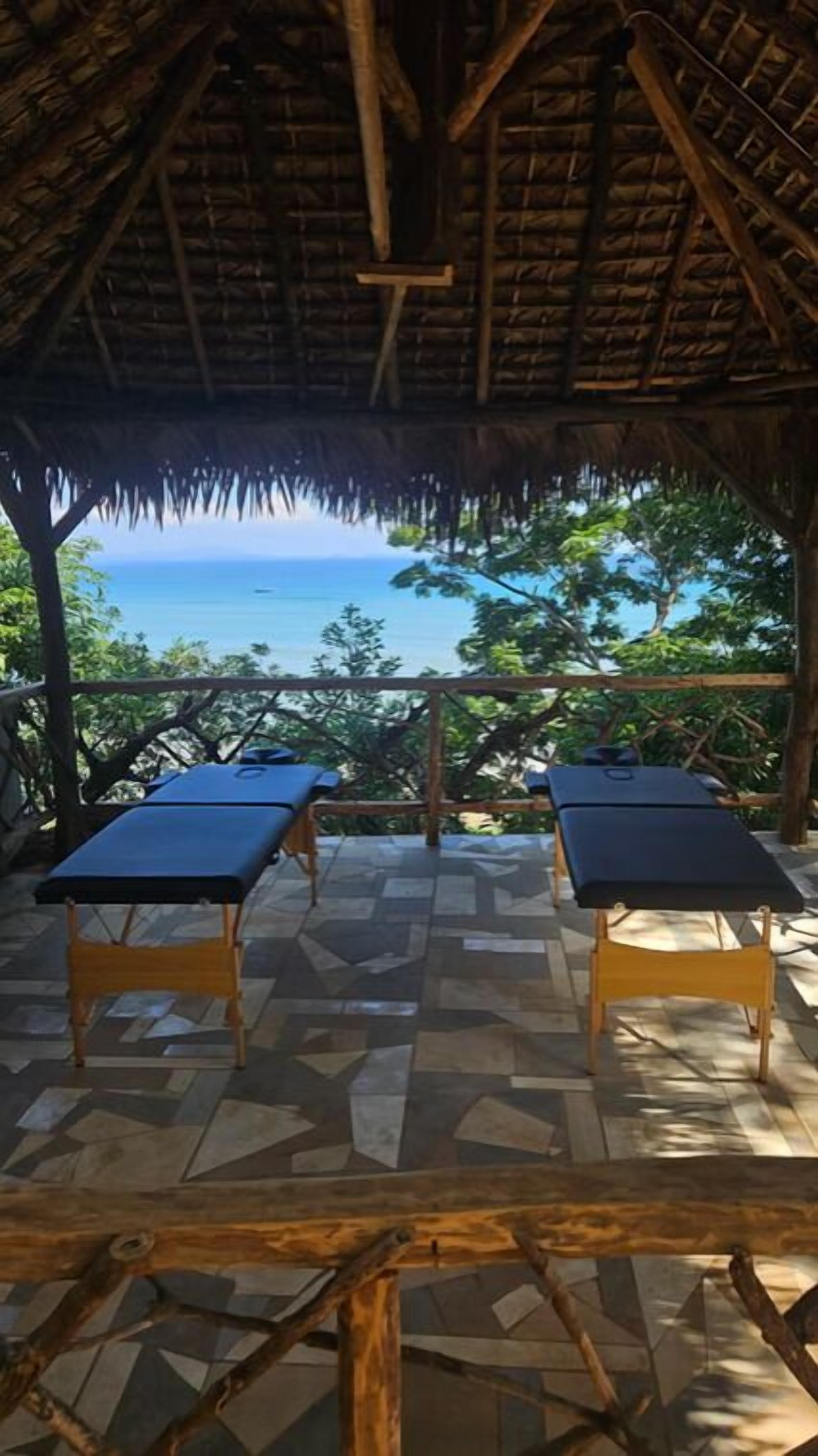 Un espace extérieur abrité comprenant deux tables de massage, un toit de chaume et une vue sur la mer.