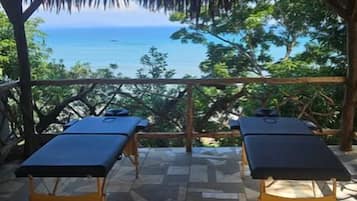 Un espace extérieur abrité comprenant deux tables de massage, un toit de chaume et une vue sur la mer.