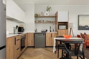 A modern kitchen with wooden cabinets, a dishwasher, and a dining area with a table and chairs. - Park Place | Lush Apartment | Balcony and Lake Views (Cape Town)