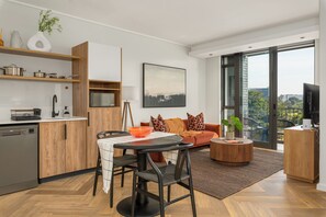 Une cuisine moderne avec des meubles en bois, une table à manger avec des chaises, un canapé avec des coussins, une table basse et une grande fenêtre donnant sur des immeubles.