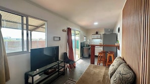 A living room with a TV, a sofa, a dining area with stools, and a kitchen in the background.