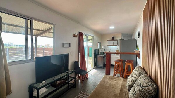 A living room with a TV, a sofa, a dining area with stools, and a kitchen in the background.