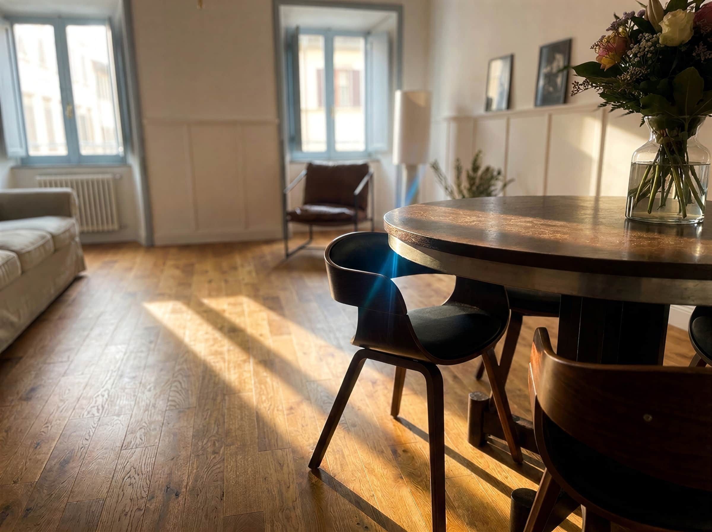 Un coin repas avec une table ronde et des chaises, un vase rempli de fleurs et une vue sur l’extérieur par une fenêtre.