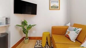 Ein Wohnzimmer mit einem wandmontierten Flachbildfernseher, einem Blumentopf, einem gelben Hocker, einer Couch mit gemustertem Kissen und einem gerahmten Bild an der Wand.