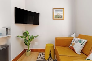 A living room with a flat-screen TV mounted on the wall, a potted plant, a yellow ottoman, a sofa with a patterned cushion, and a framed picture on the wall. - Charlie and the Chairman | Vibrant City Escape (Cape Town)