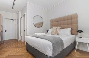A modern bedroom with a large bed, a bedside table, a nightstand, a round mirror, and a door leading to another room. - Stylish Apartment | Balcony and Pool (Cape Town)