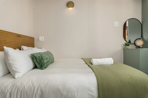 Ein ordentlich bezogenes Bett mit Kissen und Decke, ein Nachttisch mit einer Uhr und eine an der Wand befestigte Lampe.