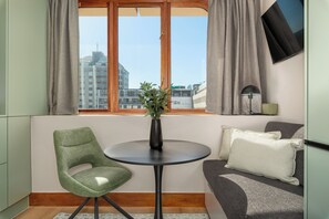 Ein kleiner Wohnbereich mit rundem Tisch, einem grünen Sessel, einer Couch mit Kissen, einer Topfpflanze und einem Fenster mit Vorhängen.