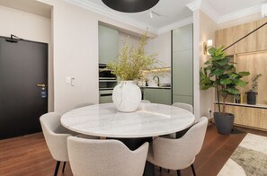 Un espace repas moderne avec une table ronde en marbre, quatre chaises au revêtement texturé, un vase blanc orné de verdure et une cuisine en arrière-plan.