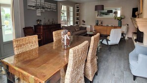 A dining area with a wooden table and wicker chairs, a large pendant light, and a view of the living room in the background. - La Zénitude***, a house located 150 m from the Bassin—relaxation guaranteed (Lège-Cap-Ferret)