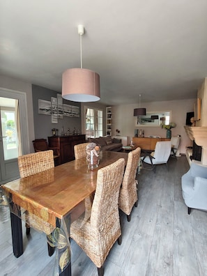 A dining area with a wooden table and wicker chairs, a large pendant light, and a view of the living room in the background. - La Zénitude***, a house located 150 m from the Bassin—relaxation guaranteed (Lège-Cap-Ferret)