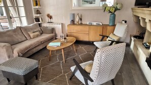 A living room with a sofa, armchair, coffee table, and patterned ottoman. - La Zénitude***, a house located 150 m from the Bassin—relaxation guaranteed (Lège-Cap-Ferret)