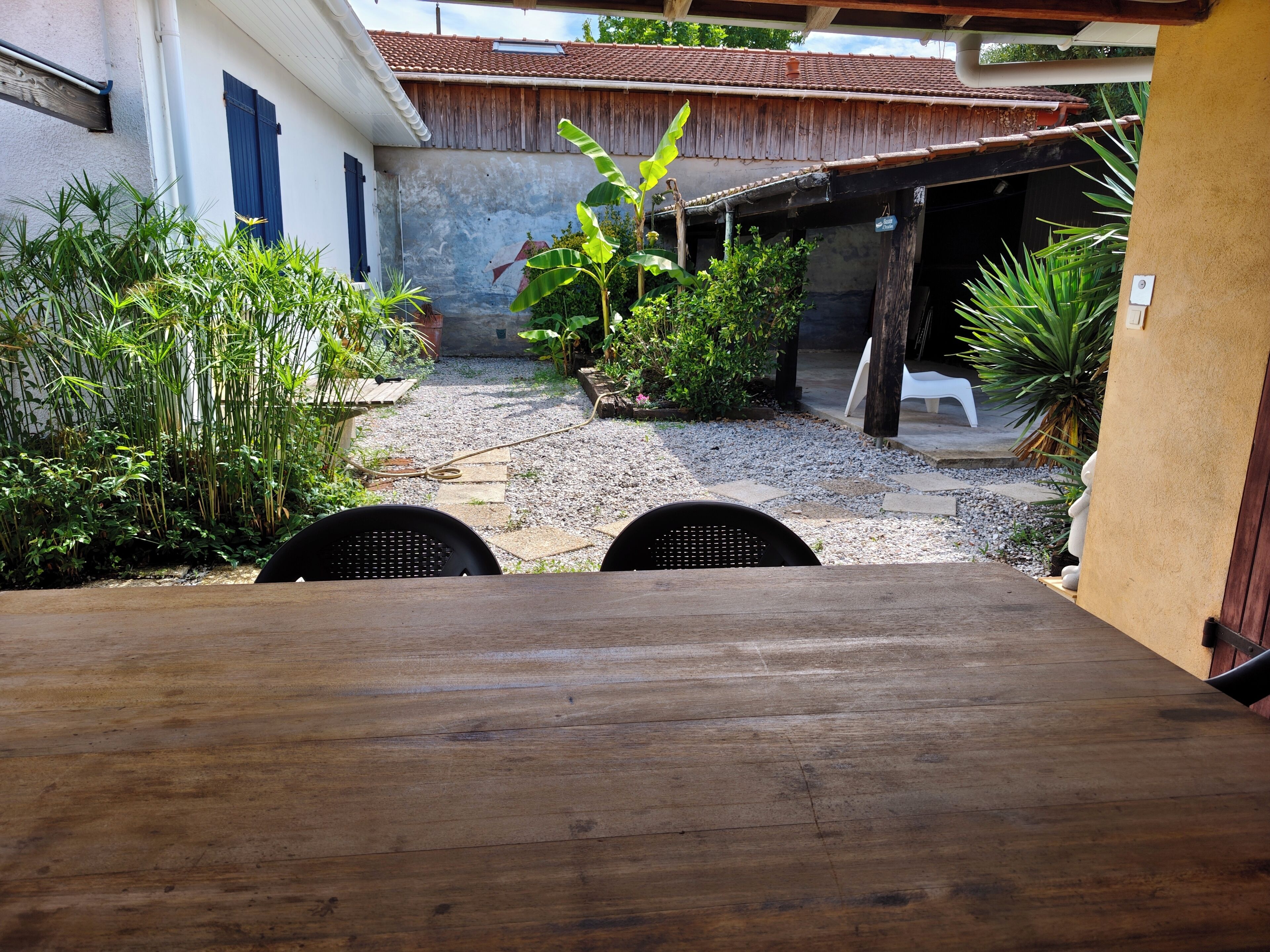 Une cour avec une table et des chaises en bois, entourée de différentes plantes et d’un bâtiment doté d’un toit en tuiles.