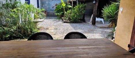 A courtyard with a wooden table and chairs, surrounded by various plants and a building with a tiled roof.