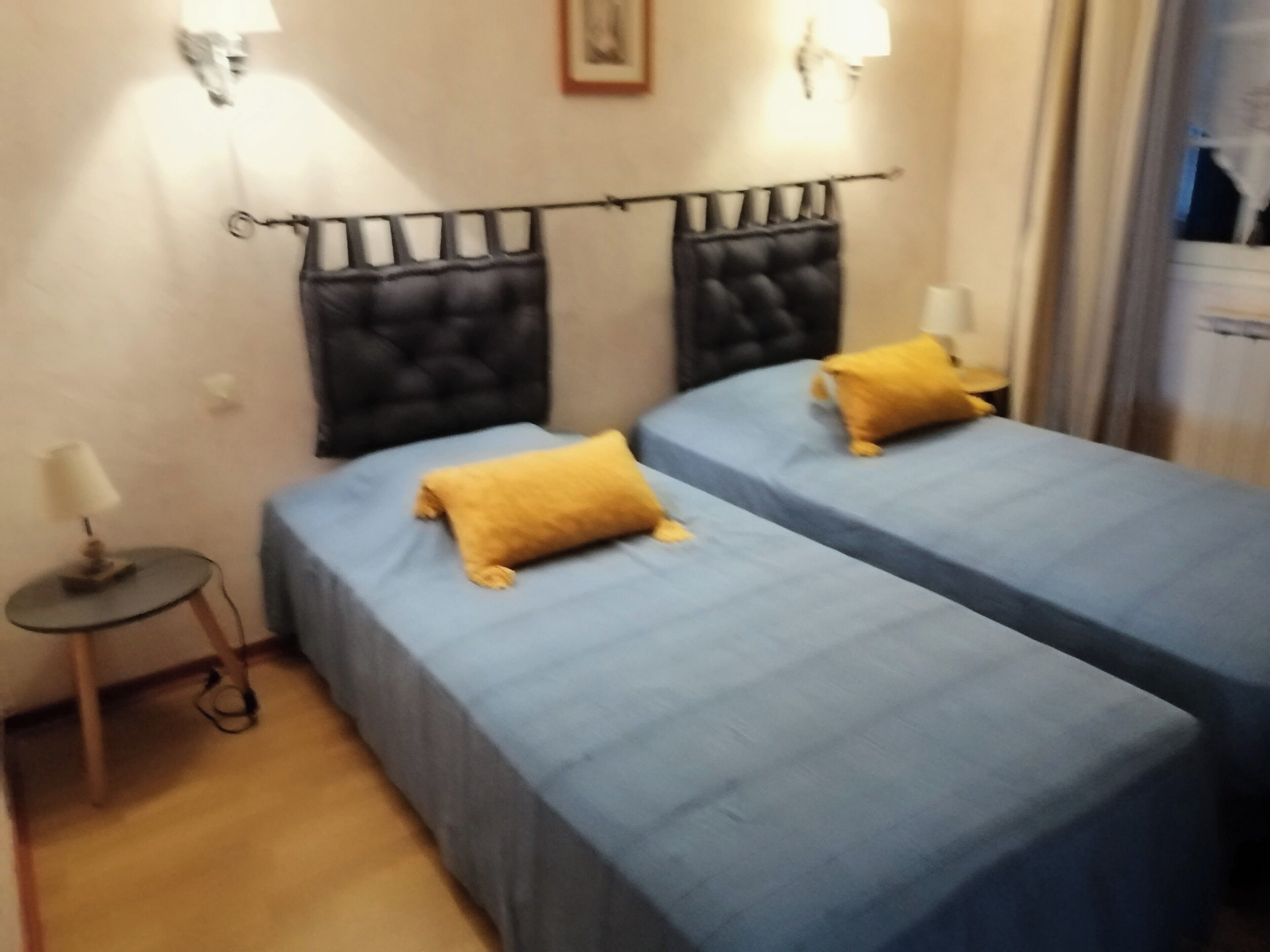 A hotel room with two beds, each with a blue bedspread and a yellow pillow. There is a small table with a lamp on the left side.