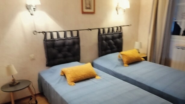 A hotel room with two beds, each with a blue bedspread and a yellow pillow. There is a small table with a lamp on the left side. - La Zénitude***, a house located 150 m from the Bassin—relaxation guaranteed (Lège-Cap-Ferret)