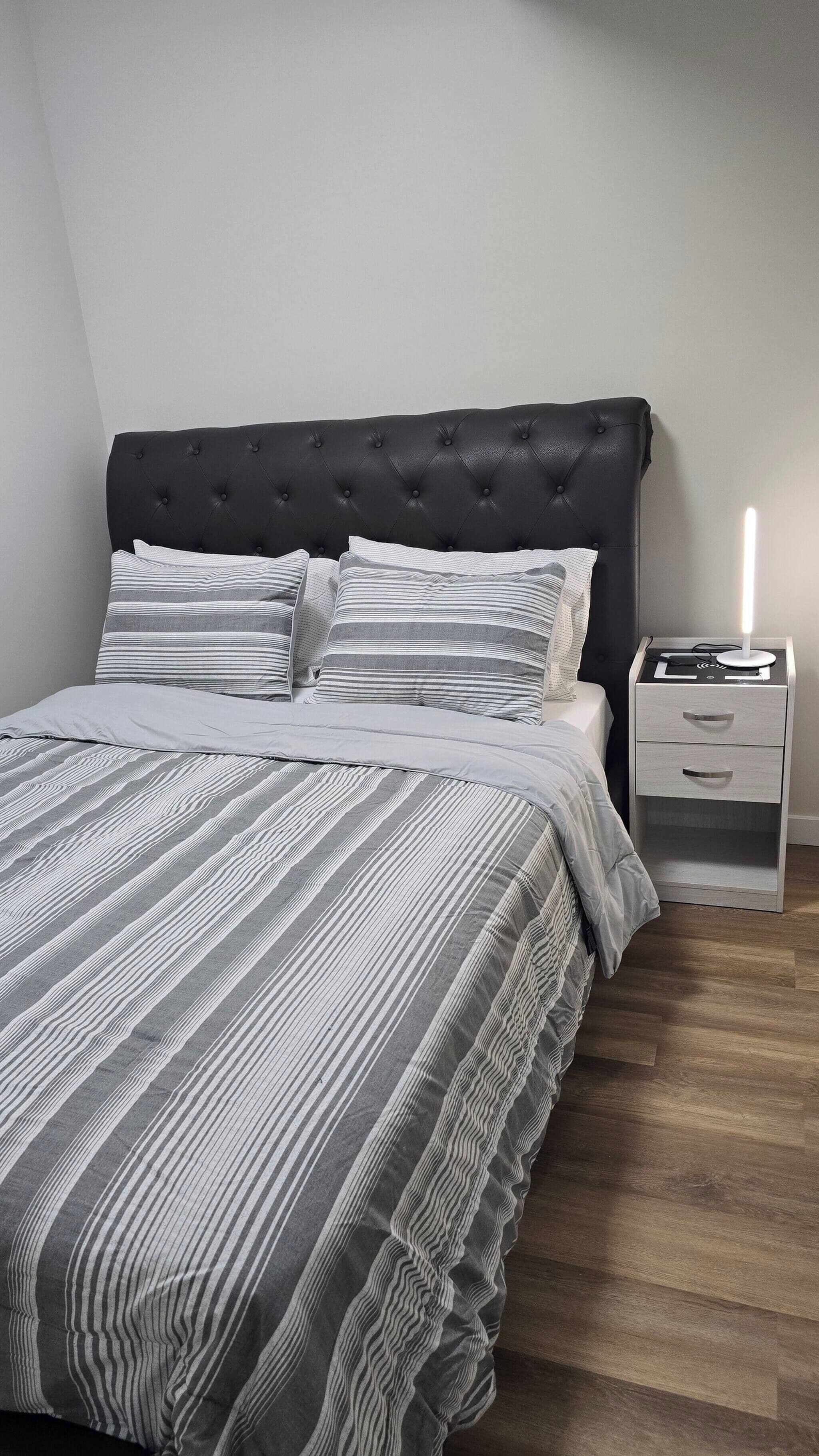 A bedroom with a tufted headboard, striped bedding, a bedside table with drawers, and a nightstand with a lamp.