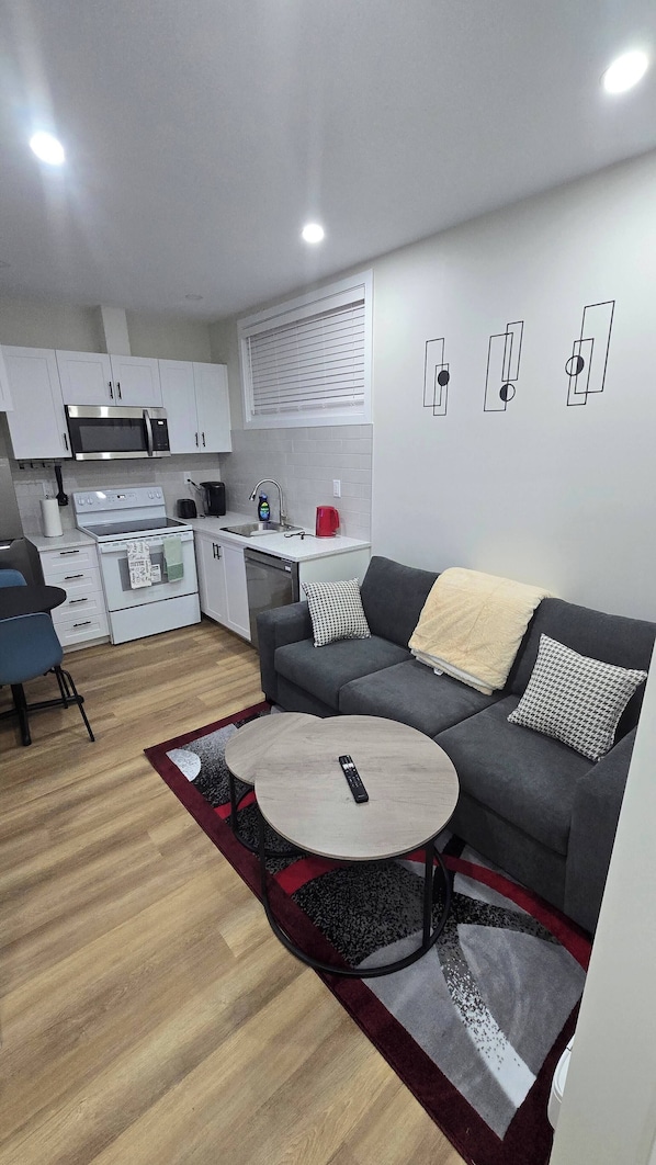 Un espacio habitable moderno con un sofá gris, una mesa redonda de centro y una cocina con armarios y electrodomésticos blancos.