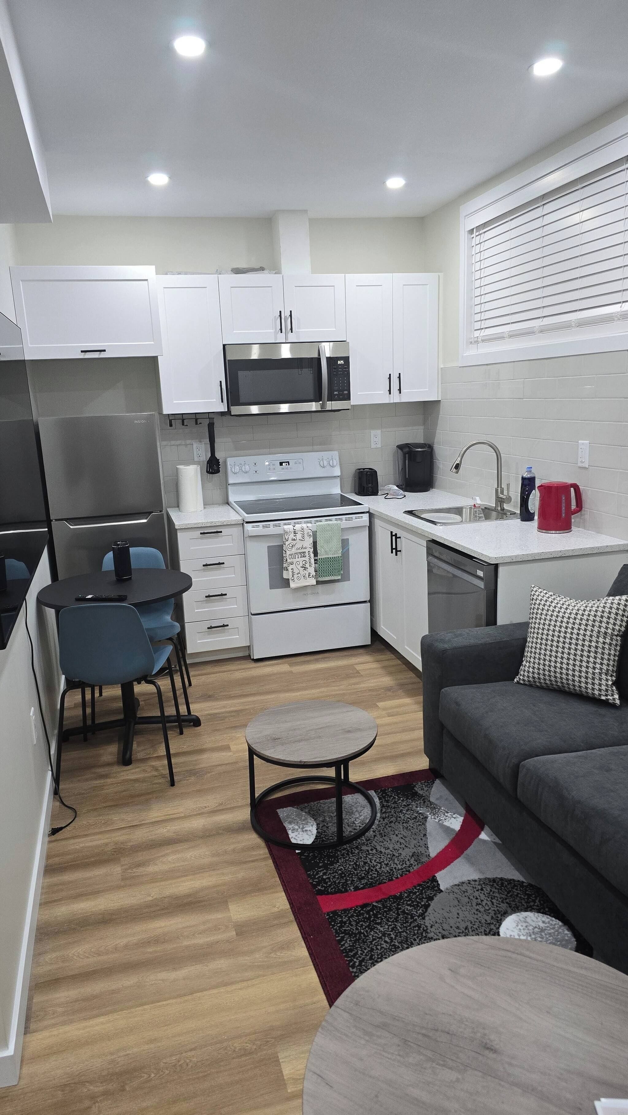 A compact kitchen with white cabinets, a microwave, a stove, and a sink. There is a small dining area with a table and chairs, and a sofa with a throw pillow.