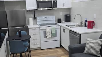 A compact kitchen with white cabinets, a microwave, a stove, and a sink. There is a small dining area with a table and chairs, and a sofa with a throw pillow.