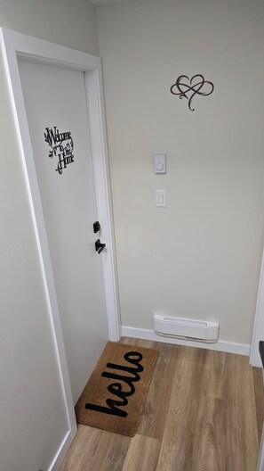 A hallway with a white door, a 'Welcome to Our Home' sign, a 'hello' doormat, a thermostat, and a white radiator.