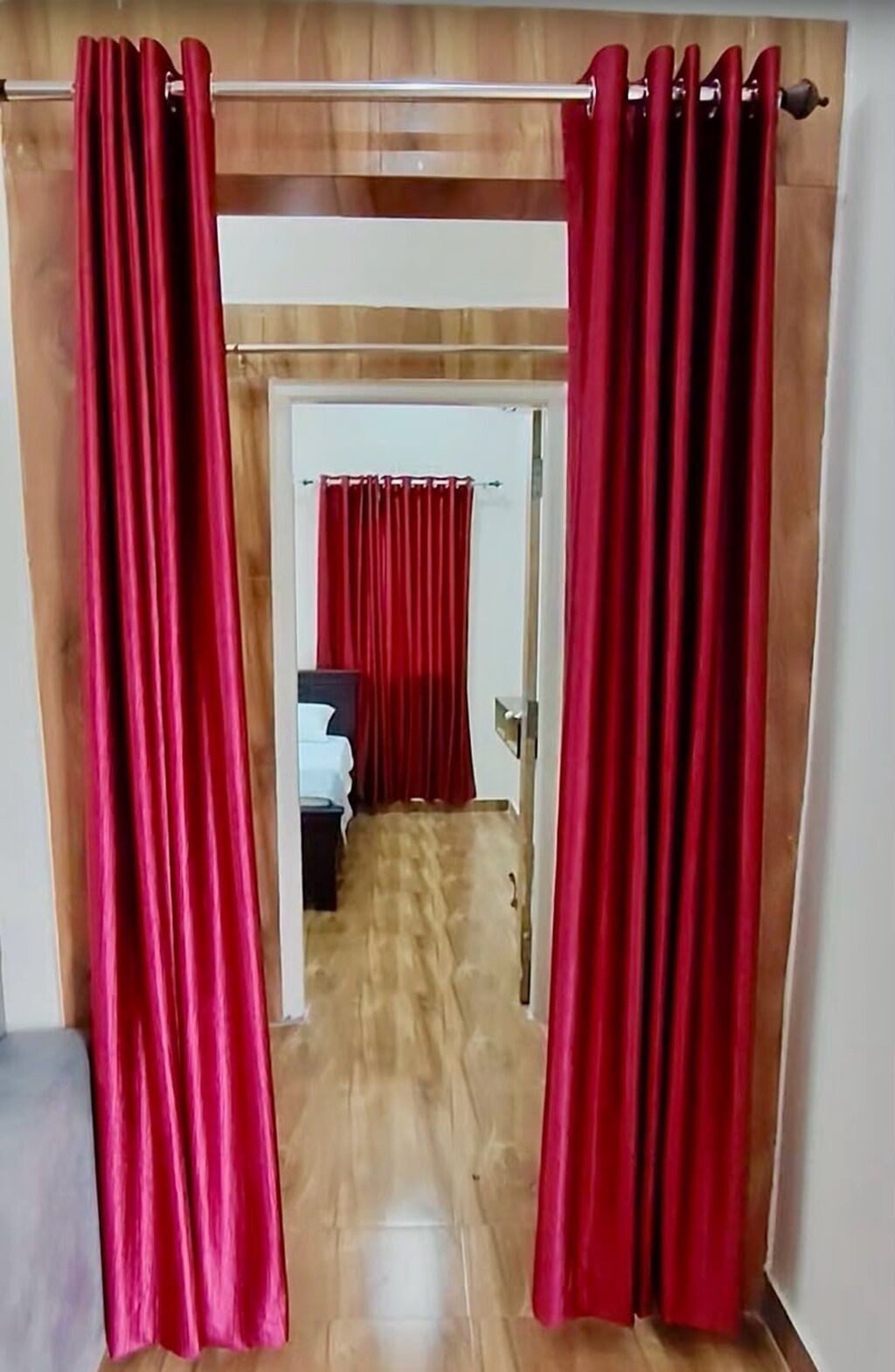 Um corredor com piso de madeira que leva a um quarto com uma cama e uma mesinha de cabeceira. Cortinas vermelhas estão fechadas na entrada.