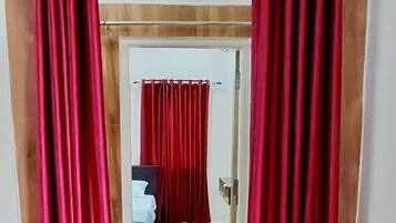 Um corredor com piso de madeira que leva a um quarto com uma cama e uma mesinha de cabeceira. Cortinas vermelhas estão fechadas na entrada.