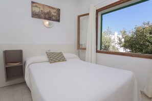 A bedroom with a bed, a bedside table, a shelf with a book, a wall-mounted light, a window with a view of buildings, and a framed picture on the wall. - Holiday home for 6 people with private pool, barbecue  Wi-Fi, air conditioning (Santanyí)