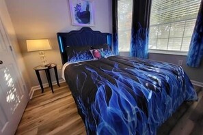 Ein Schlafzimmer mit einem Bett, einem Nachttisch, einer Lampe und einem Fenster mit Vorhängen.