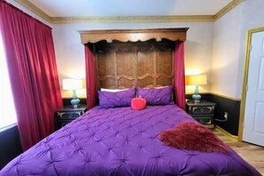 A bedroom with a large bed, two bedside tables with lamps, a wooden headboard, and red curtains. - Delightfully Villainous (Four Corners)