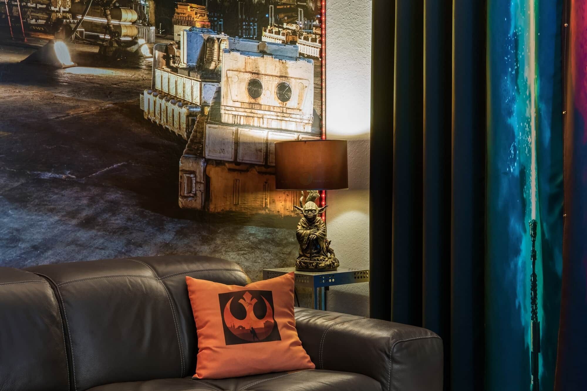 A room with a leather sofa, a pillow with a graphic design, a lamp, and a wall with a cityscape mural.