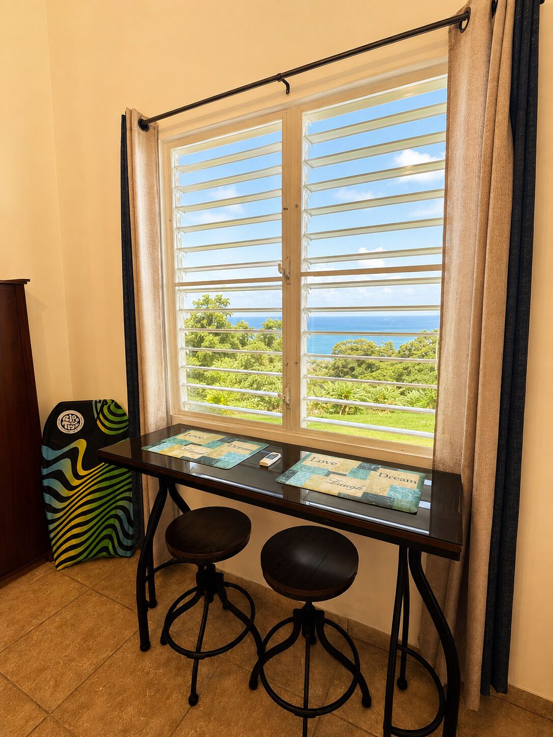 Ein Zimmer mit Fensterblick auf eine Landschaft, ein Tisch mit zwei Hocker und ein farbenfroh gestalteter Stuhl.