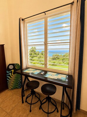 A room with a window overlooking a landscape, a table with two stools, and a chair with a colorful design.