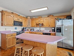 A kitchen with wooden cabinets, a stainless steel refrigerator, a pink countertop, and three wooden stools. - Decked Out in Dismal - Decks and Amenities! (Arley)