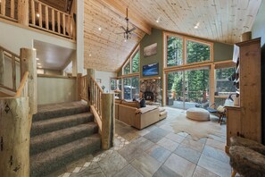 A spacious interior with a stone fireplace, a large window overlooking a forest, and a wooden staircase. - Tahoe Family Lodge | King Suite | Game Loft | Rec Center Access | Near Trails (Truckee)