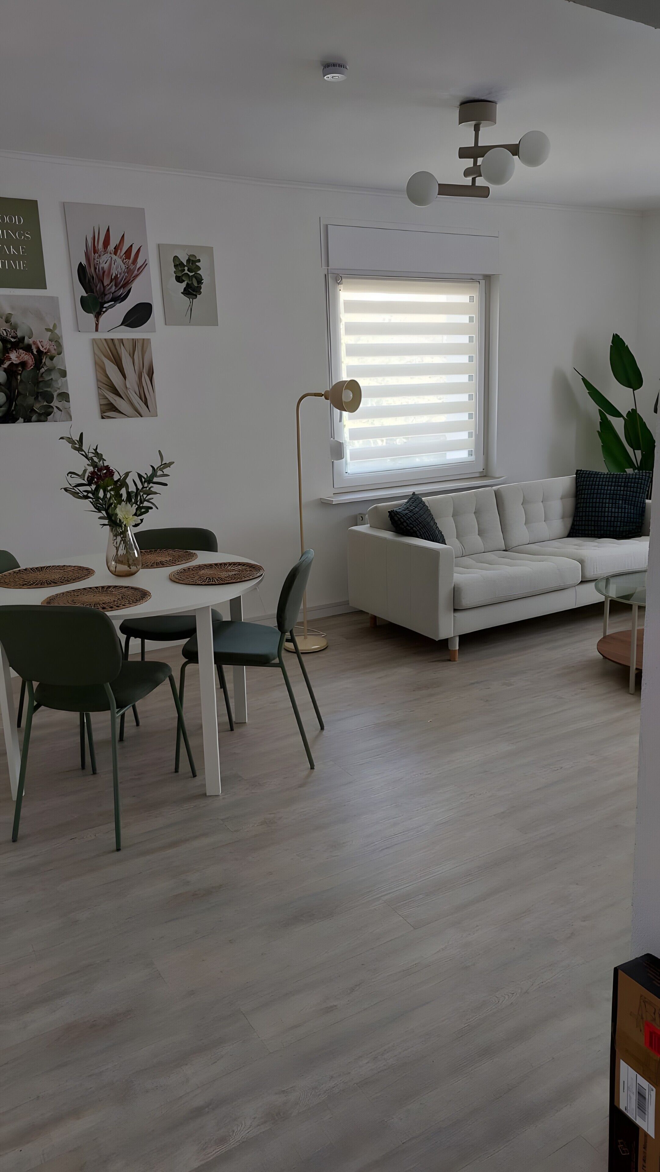 A modern living room with a white sofa, a dining table with chairs, and a large window with blinds.