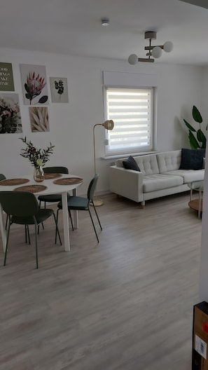 A modern living room with a white sofa, a dining table with chairs, and a large window with blinds.