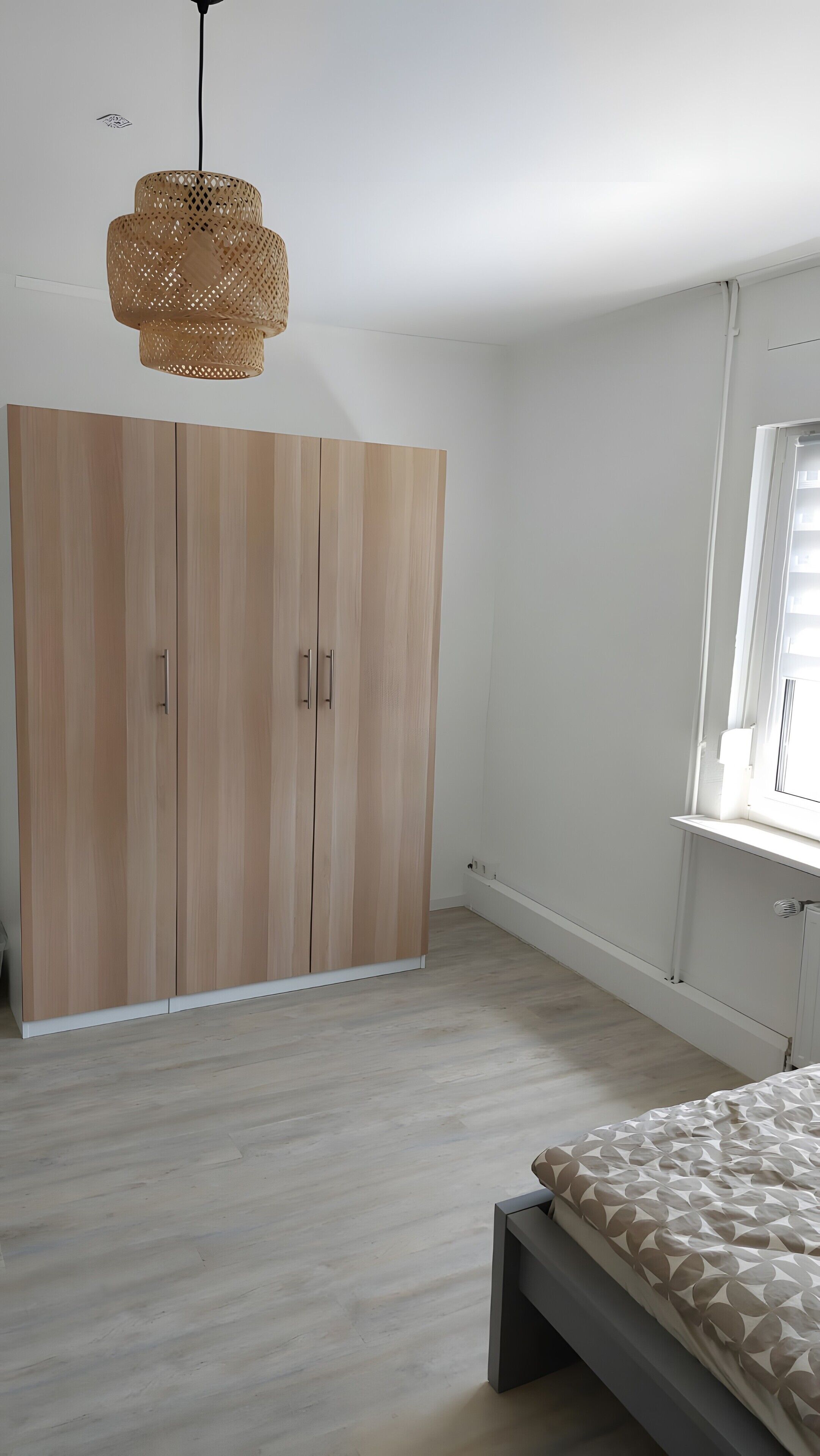 A bedroom with a wardrobe, a bed, a window, and a hanging light fixture.