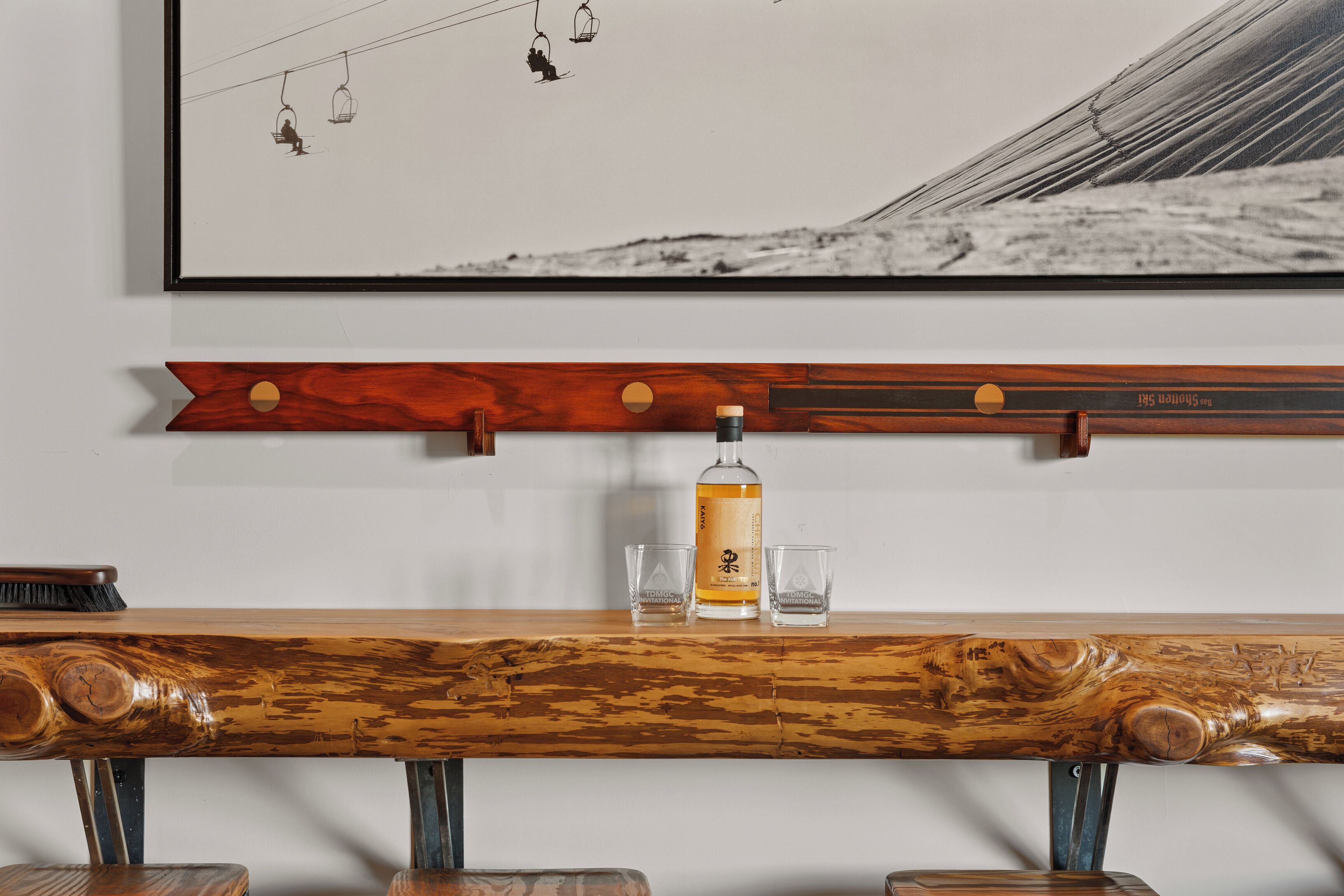 Un comptoir en bois avec une bouteille et des verres, un tableau encadré représentant une piste de ski et une étagère en bois avec une étiquette.